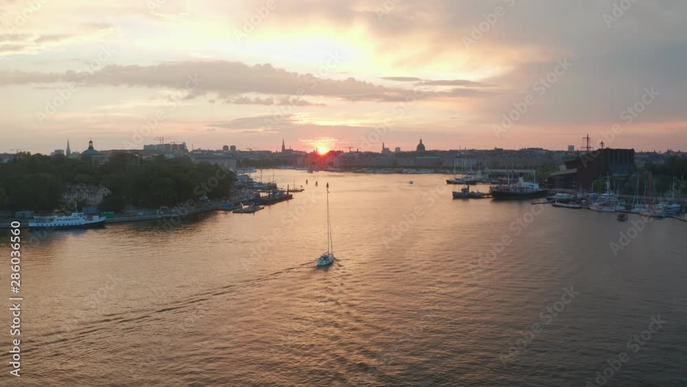 Aerial circling shot of sailboat sailing toward Stockholm city skyline during sunset.