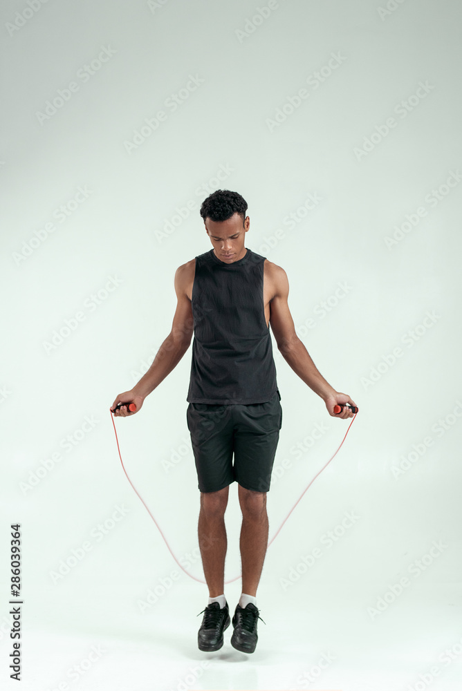 Staying fit and healthy. Full length of young african man in sport clothing skipping rope while exercising in studio against grey background