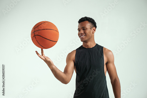 Best player. Handsome young african man in sport clothing spinning basketball ball on finger and smiling while standing against grey background