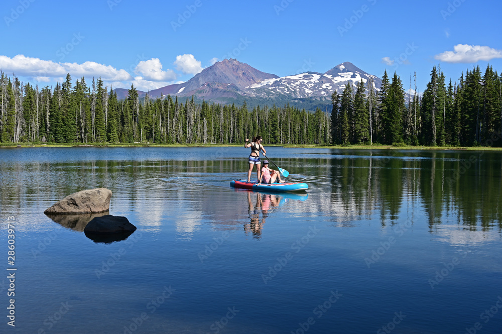 Two young women on standup paddle board on Scott Lake, Oregon with Middle and North Sisters volcanoes in background. Two young women on standup paddle board on Scott Lake, Oregon with Middle and North Sisters volcanoes in background.