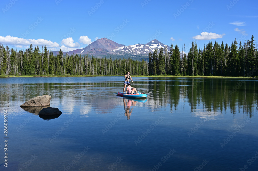 Two young women on standup paddle board on Scott Lake, Oregon with Middle and North Sisters volcanoes in background.