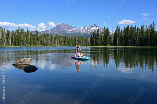 Two young women on standup paddle board on Scott Lake, Oregon with Middle and North Sisters volcanoes in background.