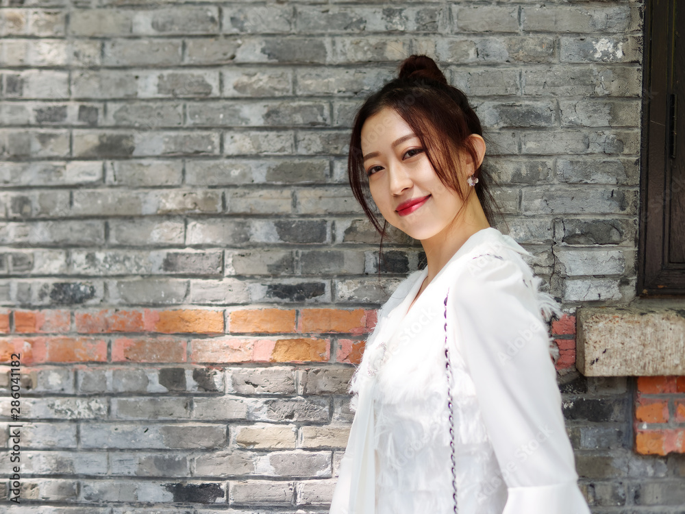 Beautiful Chinese young woman in white dress smiling at camera with brick wall background in sunny day.  Outdoor fashion portrait of glamour stylish lady.