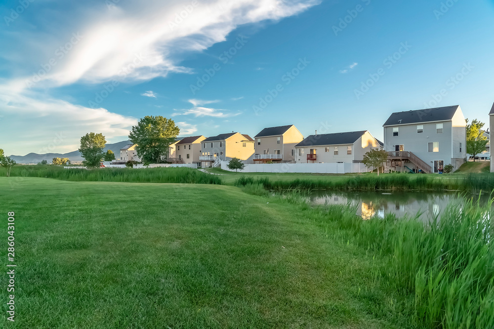 Pond and lush field in front of lovely family homes under blue sky and clouds