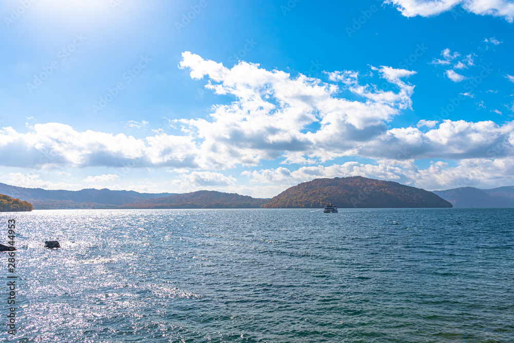 Lake Towada Sightseeing Cruises. Beautiful view, clear blue sky, white cloud, cruise ship in sunny day with autumn foliage season background. Aomori, Japan. Text in Japanese on ship 
