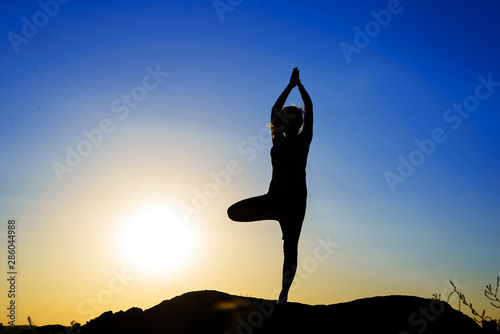 Silhouette of a young woman doing yoga on a sunset background standing on a peak of rock. Tree pose.