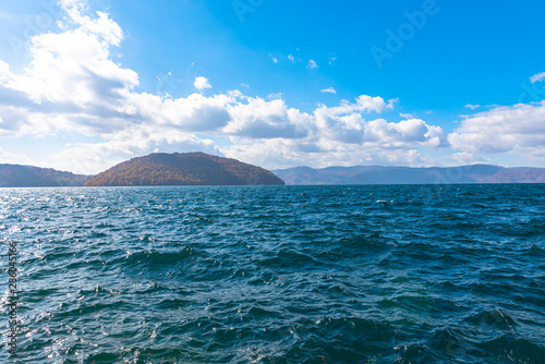 Beautiful autumn foliage scenery landscapes. Fall is full of magnificent colors. View from shore of Lake Towada, clear blue sky and water, white cloud, sunny day background. Aomori Prefecture, Japan © Shawn.ccf