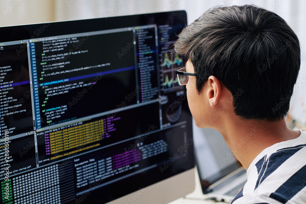 Smart teenage boy in glasses checking programming code on computer screen when sitting at his desk Smart teenage boy in glasses checking programming code on computer screen when sitting at his desk