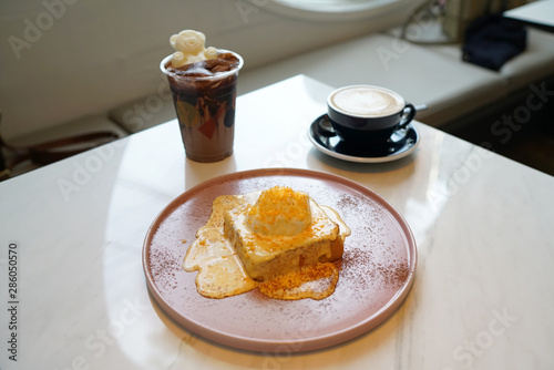 A plate of french toast (pain perdu) with salted yolk egg sauce, topped with vanilla ice cream and egg yolk flakes, served with hot latte and iced mocha coffee on marble table and blurred background.