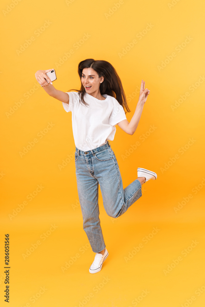 Full length image of cheerful brunette woman with long hair laughing while taking selfie photo on smartphone Full length image of cheerful brunette woman with long hair laughing while taking selfie photo on smartphone