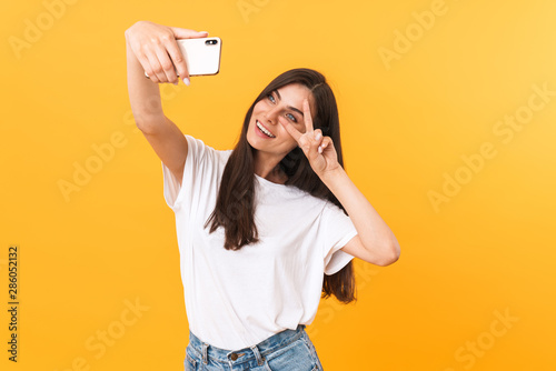 Image of pretty brunette woman showing peace sign at camera while taking selfie photo on cellphone
