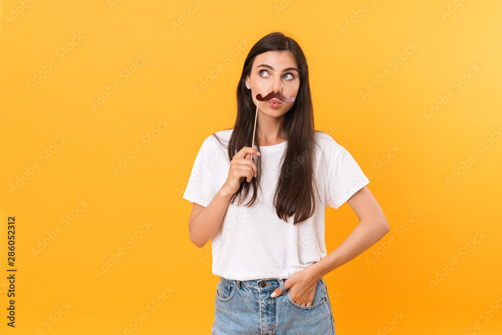 Image of charming brunette woman wearing basic clothes having fun and holding paper mustache Image of charming brunette woman wearing basic clothes having fun and holding paper mustache