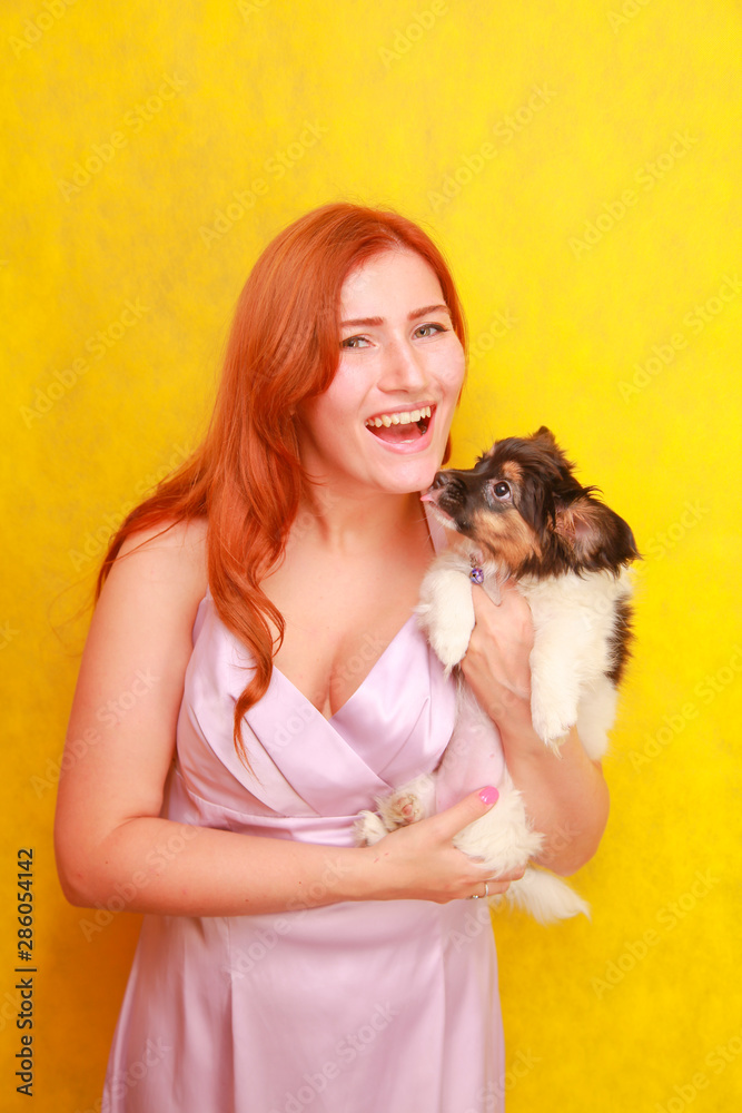 Relaxed red-haired girl embracing puppy on yellow background. Studio portrait of white appealing woman chilling with dog.