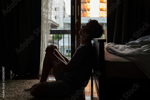 Wide shot young adult asian loneliness sad man sitting on the floor in bedroom leaning on bed and cry. Broken heart despair man sitting on the floor listening music from headphone alone.