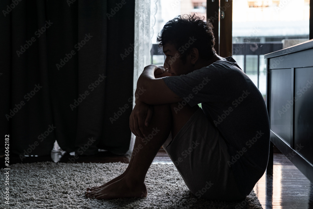 Loneliness young asian man sitting on bedroom floor alone and cry. Depression sad guy sitting hugging knees with looking away distraught and think about the problem. - obrazy, fototapety, plakaty 