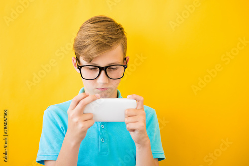 Handsome boy in glasses plays tablet on yellow background.. The concept of poor eyesight, harm of gadgets, myopia photo