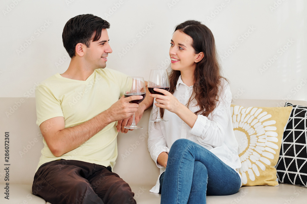 Young Caucasian couple sitting on sofa and drinking red wine when discussing news Young Caucasian couple sitting on sofa and drinking red wine when discussing news