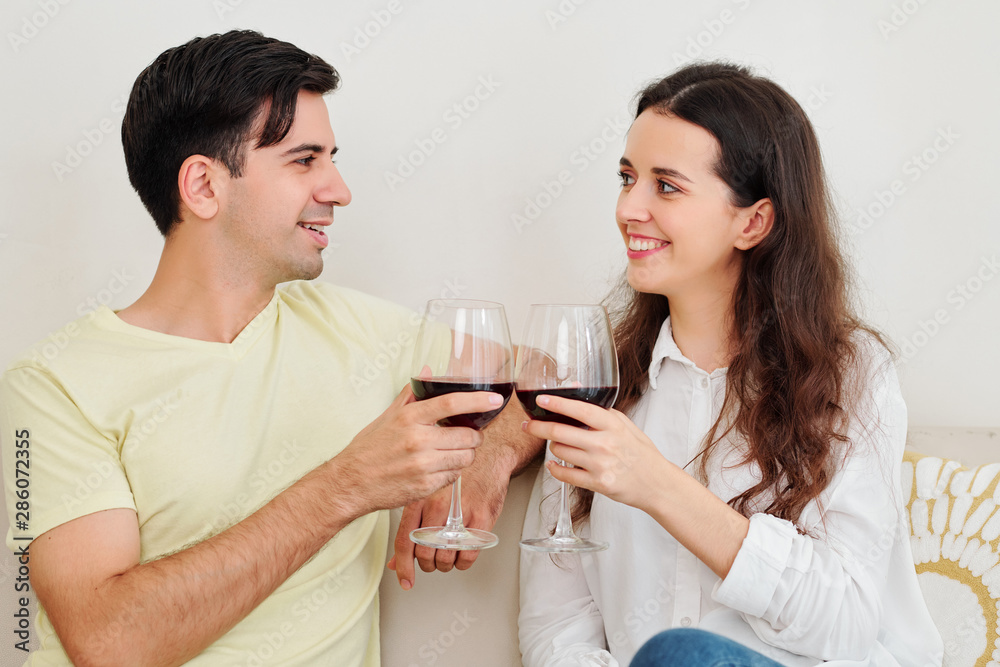 Smiling boyfriend and girlfriend toasting with wine glasses and looking at each other when having date at home Smiling boyfriend and girlfriend toasting with wine glasses and looking at each other when having date at home