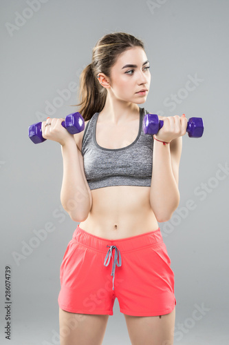 Athletic woman doing exercise for arms muscular fitness model working out with dumbbells on grey background. Strength and motivation