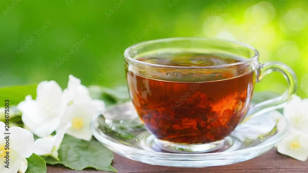 Glass cup with hot, steaming tea and jasmine blossoms in the background of the garden. Stirring tea with a teaspoon. Food conc Glass cup with hot, steaming tea and jasmine blossoms in the background of the garden. Stirring tea with a teaspoon. Food conc