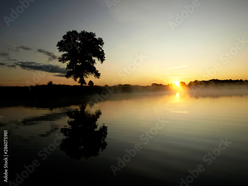Fototapeta Naklejka Na Ścianę i Meble -  foggy blured orange sunrise at a river, the burning sky is reflected in calm water , dark tree silhouettes, Salaca river, Burtnieks lake, Latvia