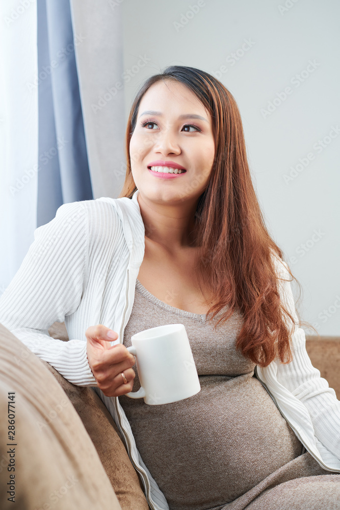 Portrait of lovely pensive young Asian woman sitting on sofa with cup of tea and looking through window Portrait of lovely pensive young Asian woman sitting on sofa with cup of tea and looking through window