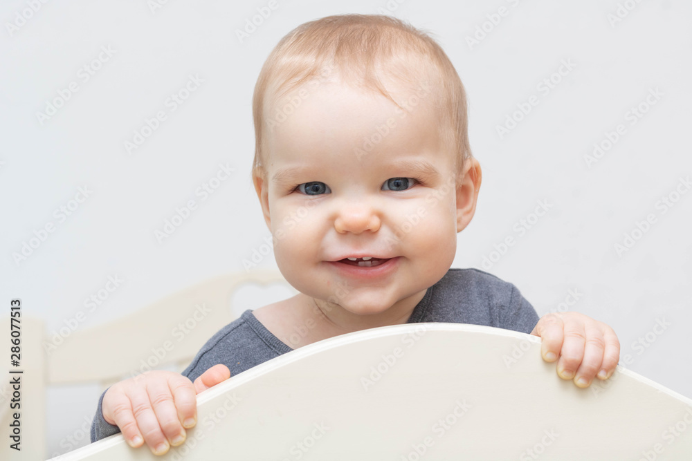 Portrait european caucasian happy smiling baby toddler with blue eyes on a white background looking at the camera holding on to the bed. Age - 10 months - 1 year. Portrait european caucasian happy smiling baby toddler with blue eyes on a white background looking at the camera holding on to the bed. Age - 10 months - 1 year.