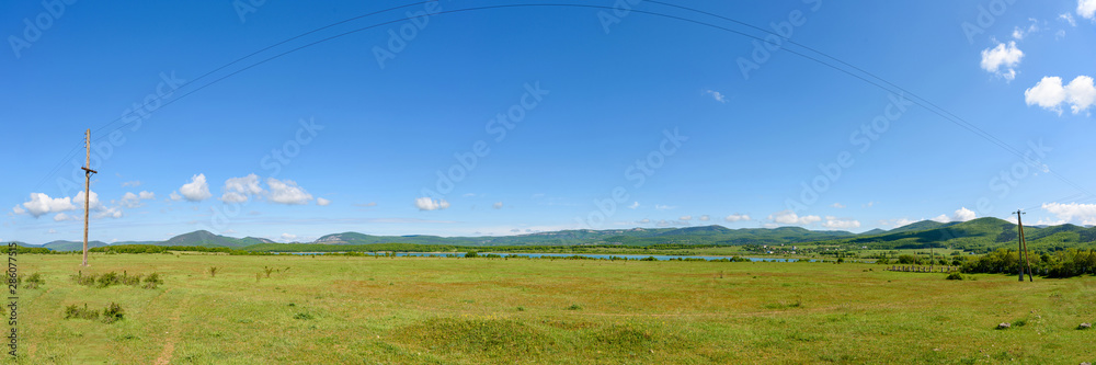 An endless field with green grass on a bright sunny day with clouds in the sky and cows perched far away near the lake.