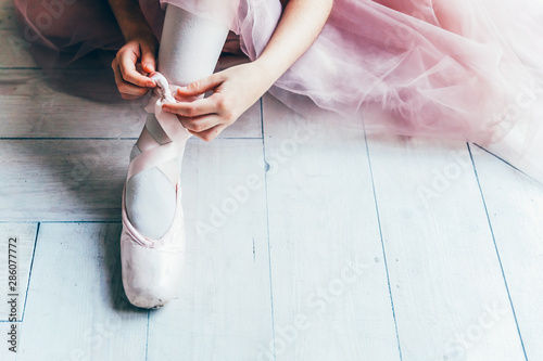 Hands of ballerina in pink tutu skirt puts on pointe shoes on leg in white light hall. Young classical ballet dancer girl in dance class photo