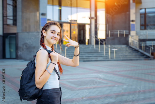 Portrait of attractive cheerful teenage girl in white t-shirt with black backpack on her shoulder. Stylish high school student makes gestures with his hands on the background of College building.
