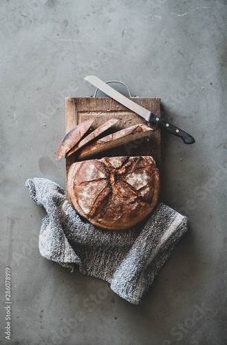 Flat-lay of freshly baked sourdough bread loaf and bread slices on rustic wooden board over grey concrete table background, top view, vertical composition photo