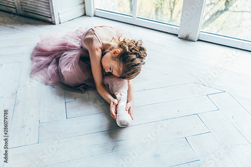 Young classical ballet dancer girl in dance class. Beautiful graceful ballerina in pink tutu skirt puts on pointe shoes near large window in white light hall photo