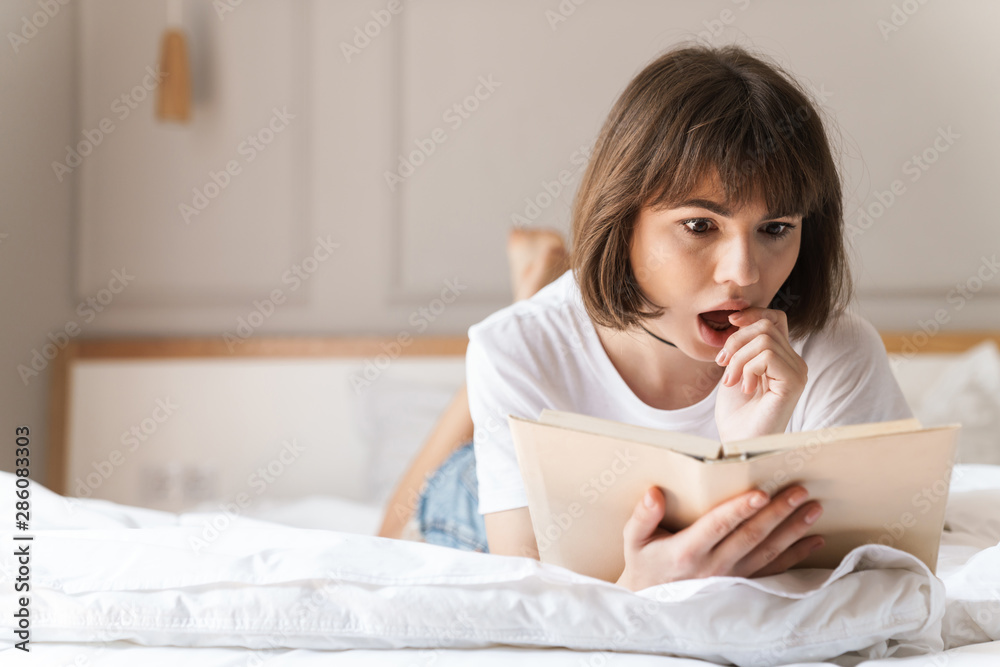 Shocked emotional young beautiful woman indoors at home lies on bed reading book. Shocked emotional young beautiful woman indoors at home lies on bed reading book.