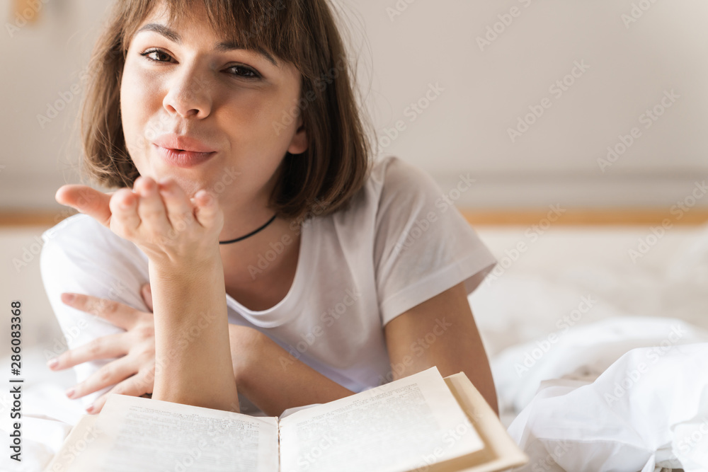 Positive optimistic young beautiful woman indoors at home lies on bed reading book blowing kisses. Positive optimistic young beautiful woman indoors at home lies on bed reading book blowing kisses.