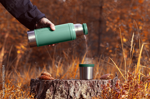 A man pours hot tea from a thermos into a mug on a stump. Autumn forest blurred