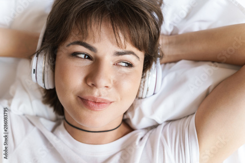 Optimistic positive pleased young beautiful woman lies indoors at home on bed listening music with headphones.