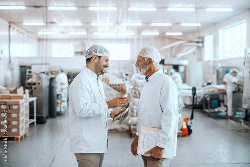 Two supervisors talking about quality of food. Younger one holding tablet while older one holding folder with charts. Both are dressed in sterile uniforms and having hairnets. Food plant interior.