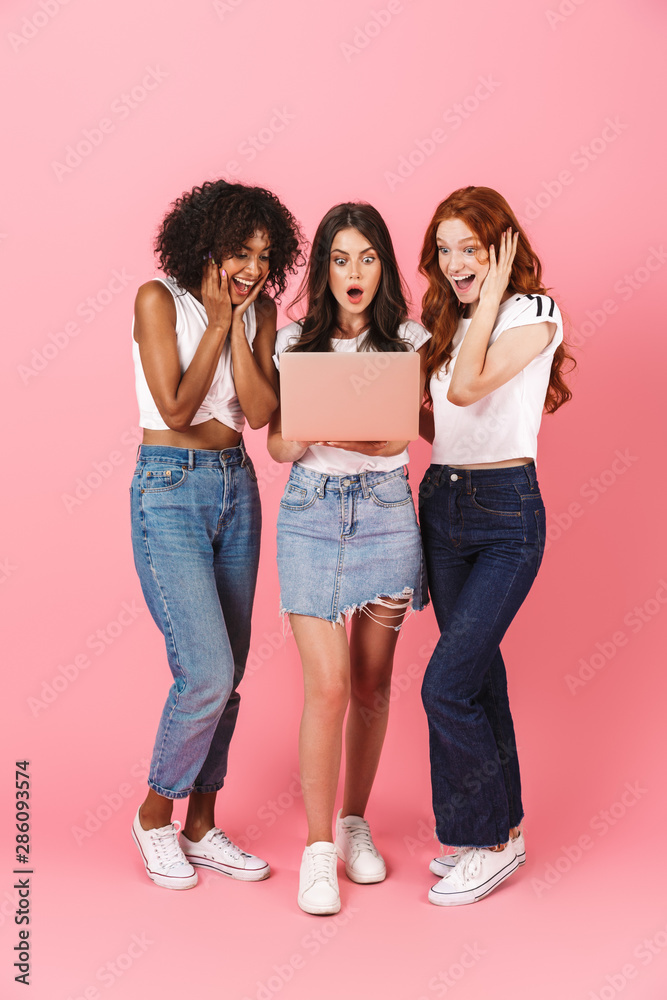 Fototapeta premium Shocked surprised emotional young three multiethnic girls friends posing isolated over pink wall background using laptop computer.