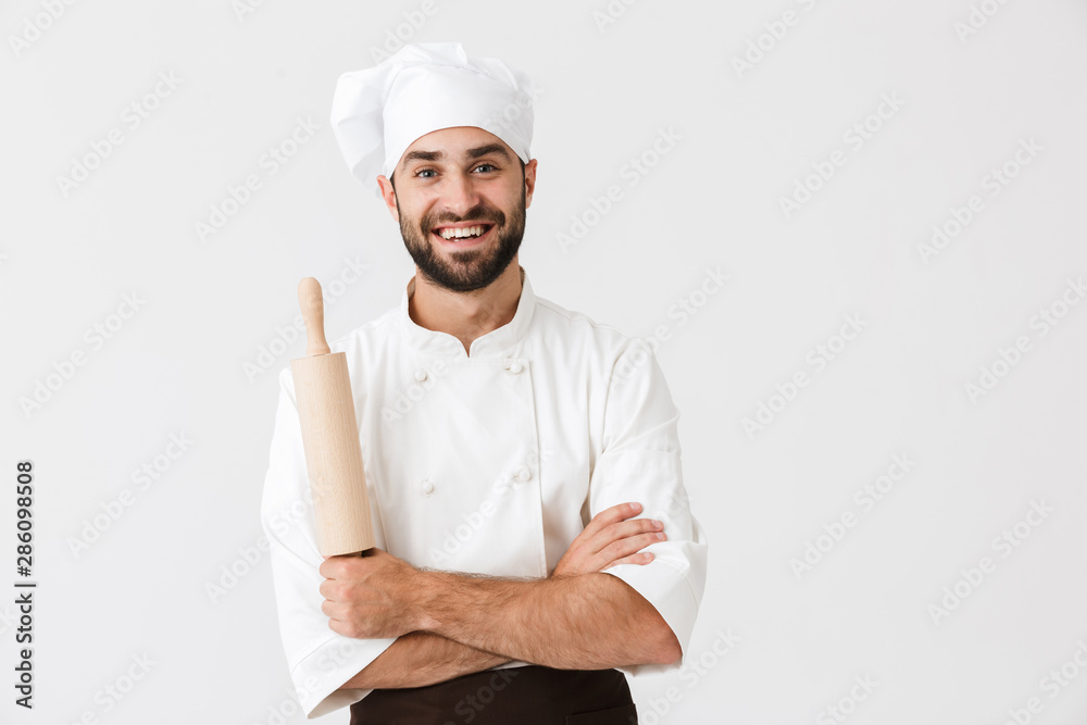 Image of happy chief man in cook uniform smiling while holding kitchen wooden rolling pin Image of happy chief man in cook uniform smiling while holding kitchen wooden rolling pin