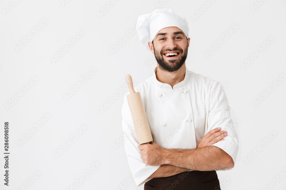 Image of young chief man in cook uniform smiling while holding kitchen wooden rolling pin Image of young chief man in cook uniform smiling while holding kitchen wooden rolling pin