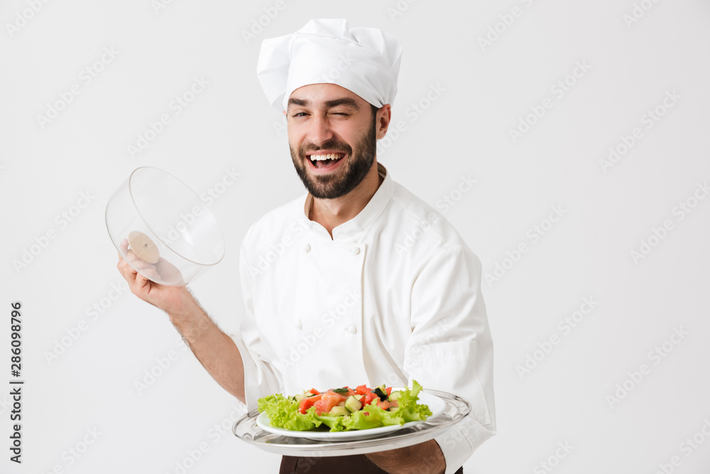 Image of handsome chief man in cook uniform smiling and holding plate with vegetable salad Image of handsome chief man in cook uniform smiling and holding plate with vegetable salad