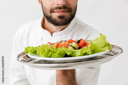 Cropped image of young chief man in cook uniform smelling dish while holding plate with vegetable salad