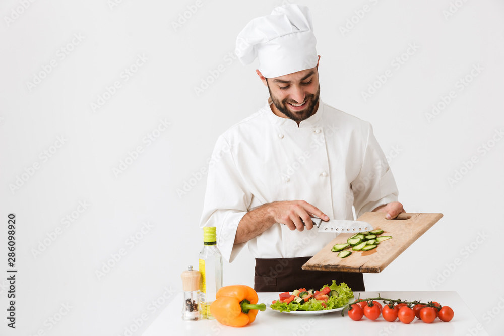Image of positive cook man in uniform smiling and cutting vegetable salad on wooden board Image of positive cook man in uniform smiling and cutting vegetable salad on wooden board