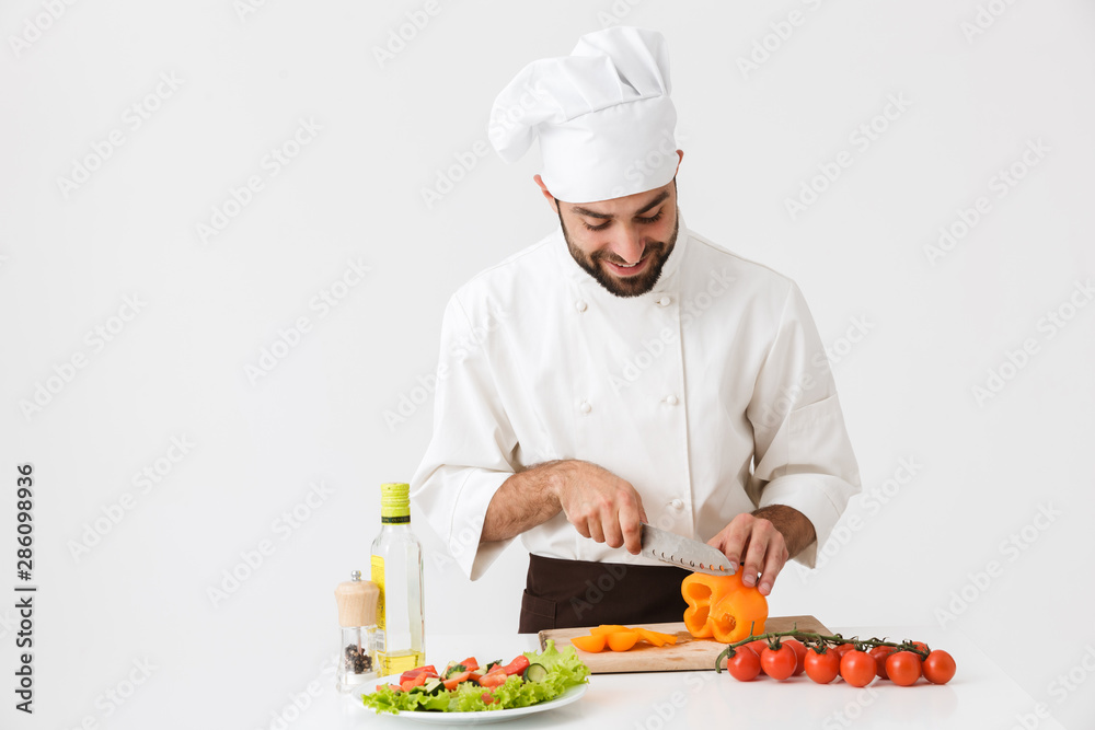 Image of caucasian cook man in uniform smiling and cutting vegetable salad on wooden board Image of caucasian cook man in uniform smiling and cutting vegetable salad on wooden board