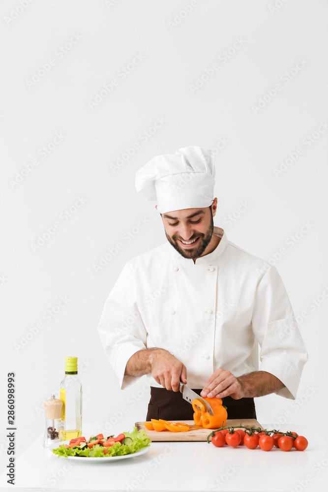 Image of pleased cook man in uniform smiling and cutting vegetable salad on wooden board Image of pleased cook man in uniform smiling and cutting vegetable salad on wooden board