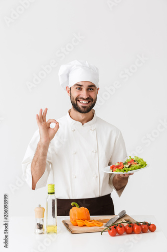Image of young cook man in uniform showing ok sign and holding plate with vegetable salad