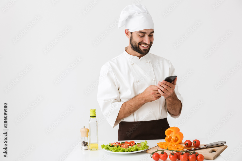 Image of pleased chef man in uniform smiling and holding smartphone while cooking vegetable salad Image of pleased chef man in uniform smiling and holding smartphone while cooking vegetable salad