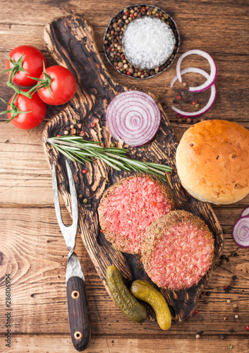 Fresh raw minced pepper beef burgers on vintage chopping board with buns onion and tomatoes on wooden background. Top view