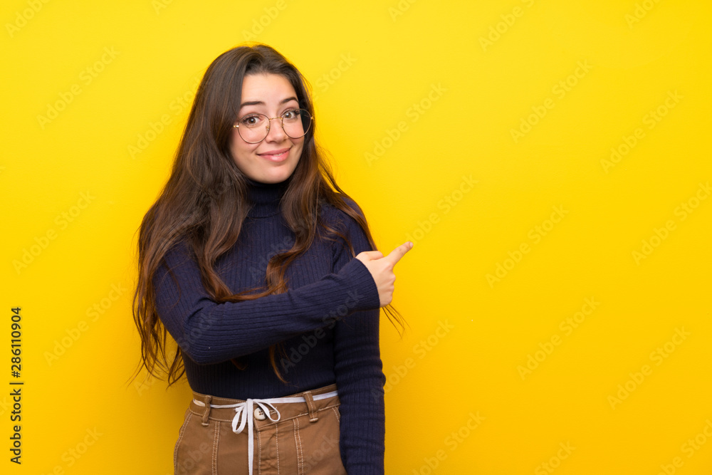 Teenager girl over isolated yellow wall pointing back Teenager girl over isolated yellow wall pointing back