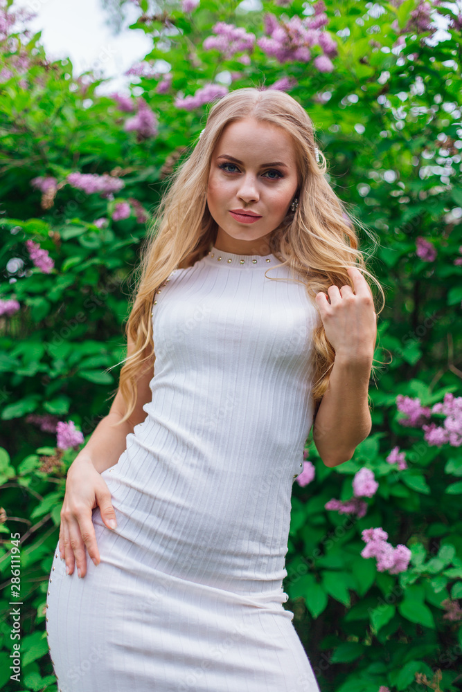 Portrait of a charming blond woman wearing beautiful white dress standing next to lilac bush. Portrait of a charming blond woman wearing beautiful white dress standing next to lilac bush.